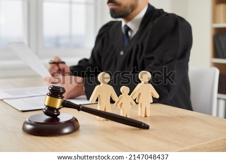 Gavel and little wooden figures of husband, wife and child up close on table in courthouse, and judge reading divorce settlement in background. Family law, court trial, parent getting custody concept