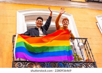 Gay boy couples with lgbt flag on balcony at home party, waving smiling, LGBT pride - Powered by Shutterstock