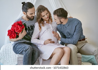 Gay couple becoming parents through surrogacy. 2 guys sitting on the bed, a pretty surrogate mom between them. One of the fathers-to-be holding roses, the other putting his hand on the pregnant belly.