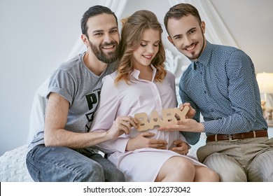 Gay couple becoming parents through surrogacy. An expectant mom sitting on the bed, loving fathers-to-be at her sides. The men smiling happily while holding lettering BABY over the pregnant belly.