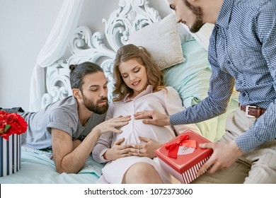 Gay couple becoming parents through surrogacy. A pretty mom-to-be hugging her tummy while lying on the bed. Two expectant dads  touching gently her pregnant belly, one of them holding a gift box.
