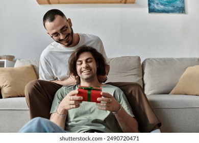 Gay couple cuddles on couch, man opens gift excitedly - Powered by Shutterstock