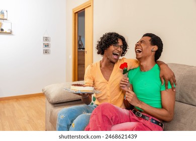 Gay couple embracing celebrating love with a rose and cake sitting together in the sofa at home - Powered by Shutterstock