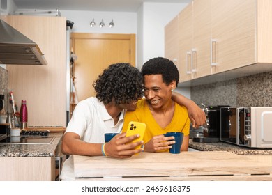 Gay couple embracing using phone and enjoying coffee cup sitting in the kitchen at home - Powered by Shutterstock