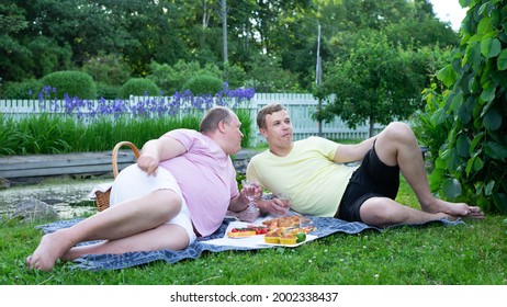 A Gay Couple Laying On The Ground In A Beautiful Garden Surrounded With Nature Having A Picnic Eating Pastries And Drinking Champagne On A Hot Summer Day Celebrating Their Anniversary.