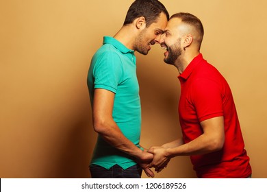 Gay Couple Love, Happy Together, Proposal, Waiting For Reply Concept. Two Young Men Holding Their Hands And Laughing Over Beige Background. White Shiny Smiles. Vibrant Colours. Copy-space. Studio Shot