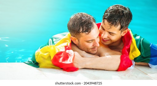 Gay Couple Relaxing In Swimming Pool With LGBT Flag. Two Young Men Enjoying Nature Outdoors, Kissing And Hugging. Young Men Romantic Family In Love. Happiness Concept