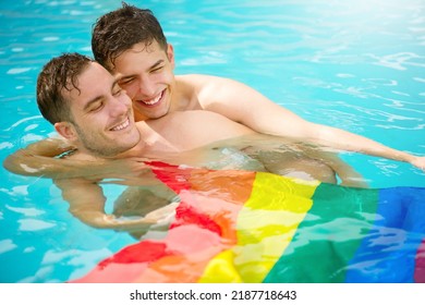 Gay Couple Relaxing In Swimming Pool With LGBT Flag. Two Young Men Enjoying Nature Outdoors, Smiling, Kissing And Hugging. Young Men Romantic Family In Love. Happiness Concept