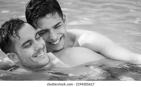 Gay Couple Relaxing In Swimming Pool With LGBT Flag. Black And White Portrait. Two Young Men Enjoying Nature Outdoors, Smiling, Kissing And Hugging. Young Men Romantic Family In Love. Happiness