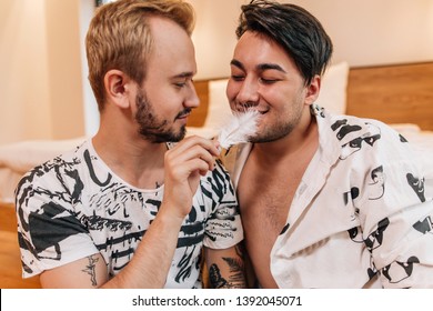 Gay couple sitting in bedroom near bed flirting with each other tickling feather lips
