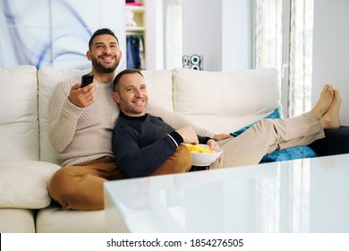 Gay couple sitting on the couch at home watching something on TV and having a snack. Homosexual relationship concept.
