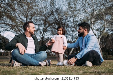 A Gay Couple Sitting In A Park Enjoying Themselves With Their Young Daughter. Modern Family.