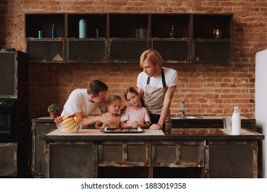 Gay Couple With Their Adopted Cute Daughters Cooking On Kitchen. Lgbt Family At Home. Portrait Of Two Handsome Men And Their Two Little Daughters Eating Cookies.