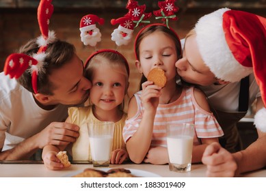 Gay Couple With Their Adopted Cute Daughters Cooking On Kitchen. Lgbt Family At Home. Portrait Of Two Handsome Men And Their Two Little Daughters Eating Cookies And Drinking Milk Wearing Santa Hat