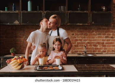 Gay Couple With Their Adopted Cute Daughters Cooking On Kitchen. Lgbt Family At Home. Portrait Of Two Handsome Men And Their Two Little Daughters Eating Cookies And Drinking Milk.