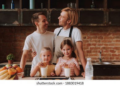 Gay Couple With Their Adopted Cute Daughters Cooking On Kitchen. Lgbt Family At Home. Portrait Of Two Handsome Men And Their Two Little Daughters Eating Cookies And Drinking Milk.