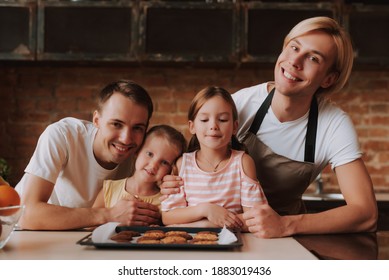 Gay Couple With Their Adopted Cute Daughters Cooking On Kitchen. Lgbt Family At Home. Portrait Of Two Handsome Men And Their Two Little Daughters Eating Cookies.