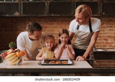 Gay Couple With Their Adopted Cute Daughters Cooking On Kitchen. Lgbt Family At Home. Portrait Of Two Handsome Men And Their Two Little Daughters Eating Cookies.