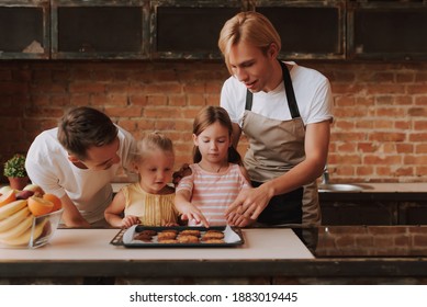 Gay Couple With Their Adopted Cute Daughters Cooking On Kitchen. Lgbt Family At Home. Portrait Of Two Handsome Men And Their Two Little Daughters Eating Cookies.