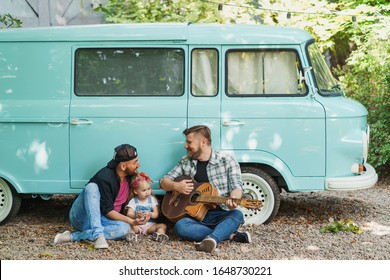 Gay Couple With Their Daughter Sitting Near A Car Van And Playin Guitar. Beautiful Moment Of Family Vacation.