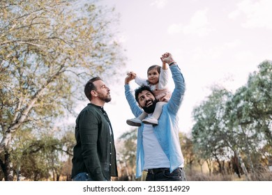 Gay Couple With Their Young Daughter Enjoying A Day In A Park. The Father Raises The Arms Of The Daughter Smiling At Her.