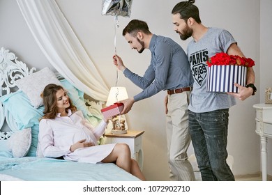 Gay Couple Waiting For Their Surrogate Baby. The Dads-to-be Pampering The Expectant Mother, Giving Her Presents And Flowers. She's Relaxing On The Bed While Taking Gifts With A Smile On Her Face.