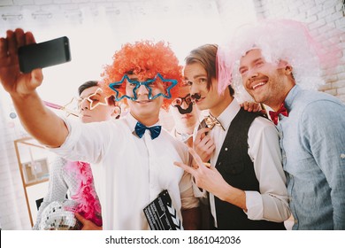 Gay guys in bow ties taking selfie on phone at party. Man with glasses and an orange wig stands and takes a photo with friends