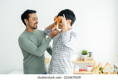 Gay LGBT Sweet Happy Asian Couple Wearing Pajamas, Smiling, Taking Care, Teasing With Piece Orange On Eyes, Eating, Healthy Fruit For Breakfast In Kitchen At Home In Morning. Lifestyle, Love Concept