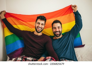 Gay Male Couple Holding Lgbt Rainbow Flag Indoors On Bed At Home - Focus On Right Man Face