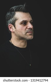 A Gay Man In Black Shirt On A Black Backdrop - Wearing A Pride Necklace - Has Salt And Pepper Hair And Stubble. 