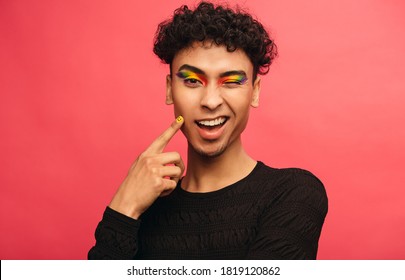 Gay man wearing multicolored shadows on the eyelids winking at camera. Happy transgender male winking an eye against red background.