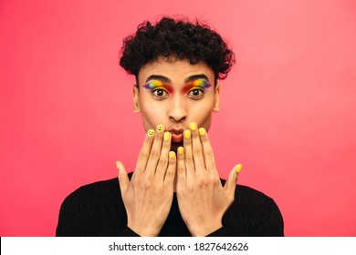 Gay man wearing rainbow colored eye shadow and smiley face on fingernail on red background. Transgender male with funky makeup.