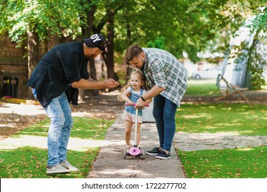 Gay Parents Teaching Their Daughter To Ride A Scooter. Young Family Spending Joyful Time In The Park. LGBT Parenting Concept.