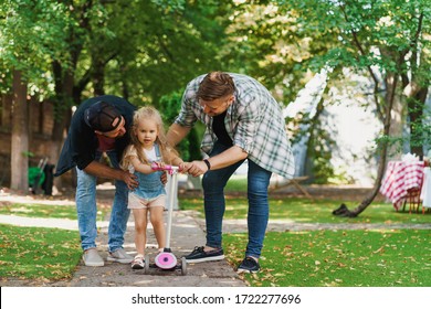 Gay Parents And Their Daughter In The Park. Careful Dads Helping The Child To Ride A Scooter For The First Time.