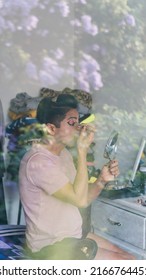 Gay Person Applying Makeup Using A Mirror And Eyeliner, Sitting On A Bed.