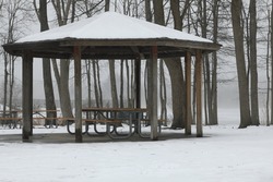 Gazebo Covered In The Snow With Picnic Bench Under The Covering. Forest And Frozen Lake In The Background. 