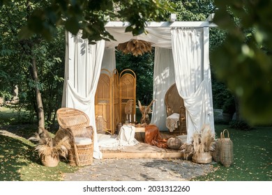 Gazebo decor at nature, outdoor design of arbour. Vintage decoration outside, natural rural background. White curtain, dry plants and wicker furniture for summer house in park.