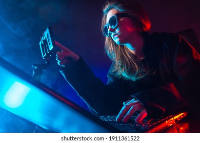 Geek woman next to a laptop. She uses a cell phone. Portrait of a geek woman in a dark room. Geek woman face is illuminated with neon light. Young girl works with a computer. She prints something