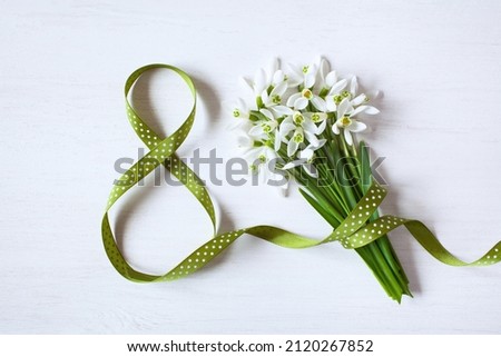 Geeting card for Women's day march 8, number eight from a green ribbon and a bouquet of snowdrops on a white wooden background