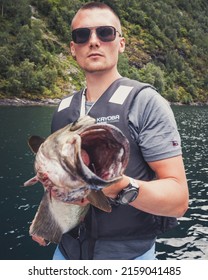 GEIRANGER, NORWAY - Aug 24, 2019: The Fisherman On A Boat In The Sea Holding In Hands Fish With Open Mouth In Geiranger , Norway In Summer