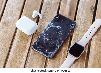 Gelendzhik, Russia, 29/07/2020: iPhone with broken back glass in cracks lies on wooden table next to headphones and Applewatch