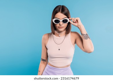 gen z fashion, brunette young woman with short hair in tank top, skirt and sunglasses posing on blue background, casual attire, stylish posing, personal style, portrait, fashionista - Powered by Shutterstock