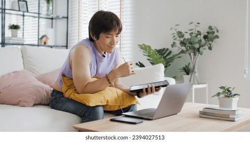 Gen Z Gay queer man non-binary sitting easy relax self study learn online happy work at home sofa. Asia people LGBT young adult guy WFH listen video call reskill upskill in MBA career job school class - Powered by Shutterstock