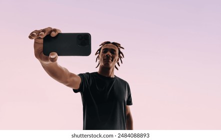 Gen Z man with dreadlocks uses a smartphone to take a selfie. The selfie camera app, and studio backdrop reflect his tech-savvy, connected lifestyle and social media presence. - Powered by Shutterstock