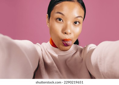 Gen Z woman taking a fun selfie while wearing bold lipstick in a studio. Young woman sticking her tongue out and looking at the camera as she takes a self-portrait. - Powered by Shutterstock
