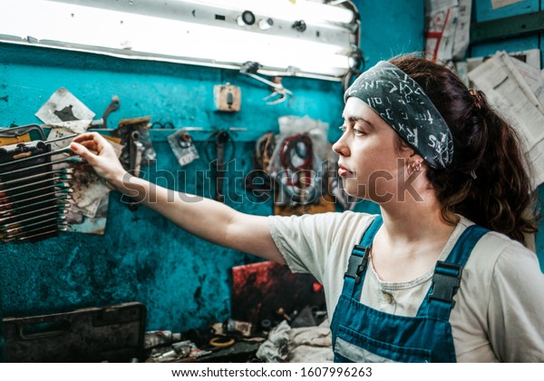 Gender equality. Portrait of a young pretty woman in\
uniform working in a workshop, who takes out a wrench from a\
toolbox on the wall. Side\
view