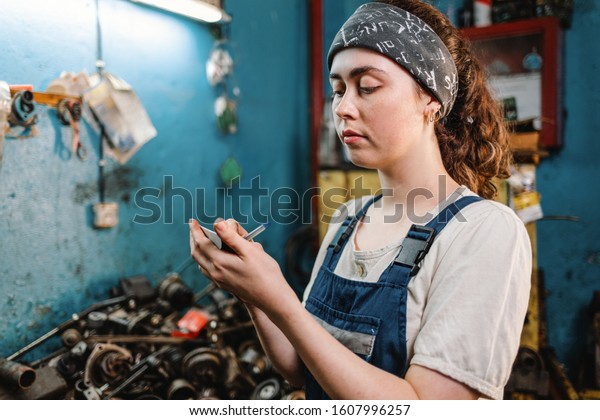 Gender equality. Portrait of a young woman in uniform,\
working in a workshop, which writes down orders on a piece of\
paper. In the background is a blue wall and a table with spare\
parts. Close up