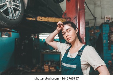 Gender Equality. A Young Brunette In Uniform Stands Near The Elevator With A Car And Wipes The Sweat From Her Forehead. In The Background Is An Auto Repair Shop