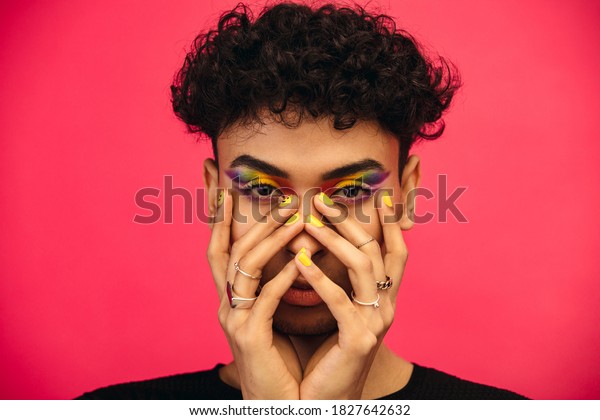 Gender fluid male wearing rainbow colored eye
shadow and smiley face on fingernail. Transgender male with funky
makeup on red
background.