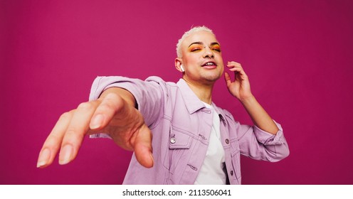 Gender non-conforming man enjoying his favourite song. Non-conforming queer man dancing while listening to music on wireless earphones. Young man standing alone against a purple background.